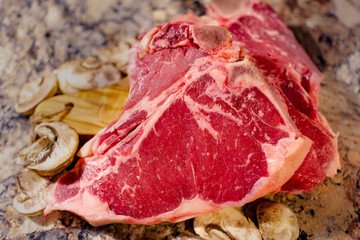 A T bone or Porterhouse raw steak on cutting board with mushrooms. Blurred foreground. Blurred background. Neutral marbled surface.