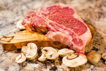 A T bone or Porterhouse raw steak on cutting board with mushrooms.  Blurred foreground.  Blurred background.  Neutral marbled surface.