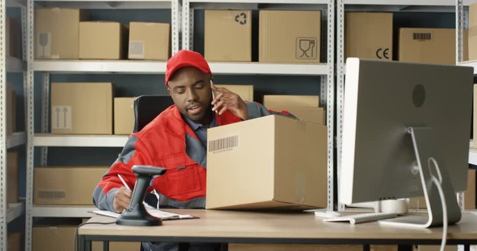 African American young male postman in uniform talking on cellphone and scanning parcel. Man registering carton box mail and filling in invoice while speaking on phone and sitting at computer.