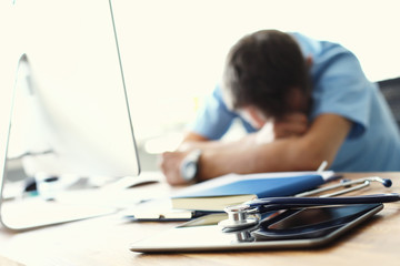 Tired man doctor sleeps lying on his hands while sitting at a computer desk. Doctor works on a computer in the clinic after a night shift.