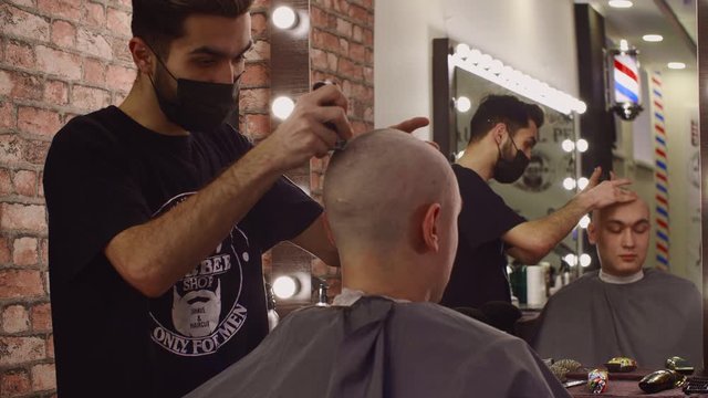 Professional hairstylist shaving bald head of client. Concentrated hairdresser in facial mask serving male client in barbershop. Male beauty concept