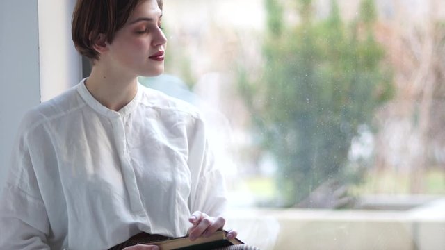 Woman is sitting at the window and reading a book. Close Indore portrait of a young girl in a vintage white blouse. Happy woman, calm weekend, meditation and self-knowledge