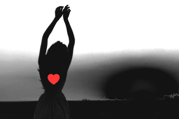 Silhouette of woman with heart and with hands up against sky on dark black background. Place for...