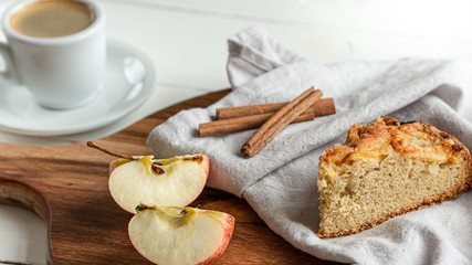 Apple pie slice lying on a grey cotton napkin. Wooden cutting board. There is cinnamon sticks, apple slices. And cup of coffee on the background. Wooden white background. 