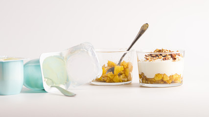 Empty blue yogurt container lying on a table with the spoon in it. Natural yogurt in a blue plastic container. Near there is a glass with some apple jam, yogurt and  pecan nuts. White background. 