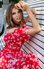 Fototapeta premium Beautiful young girl, model, brown-haired woman with long hair in a red dress sits on a bench and poses with a relaxed, languid look.