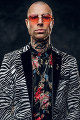 Vivid, calm, tattooed, bald male model posing in a dark studio for the photoshoot wearing fashionable custom made zebra striped style tuxedo, golden chain, rose patterned shirt and red glasses, dandy