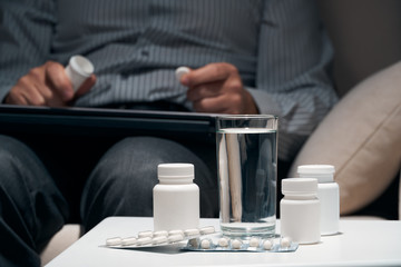 Fototapeta na wymiar Overworked businessman sitting on the sofa and taking medication. Pills and glass of water near him.