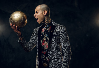 Expressive, fashionable, tattooed, bald male model posing in a studio for the photoshoot wearing fashionable custom made zebra style tuxedo, rose patterned shirt, looking on a golden soccer ball while