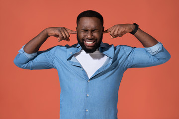 Man plugs his ears with hands for not hear anymore. Photo of african man in casual outfit on coral...