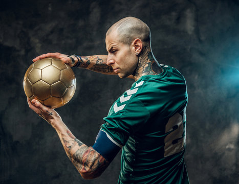 Bold and handsome soccer player posing for a photoshot in a dark studio, wearing professional sportswear, and holding a golden soccer ball in one hand, standing sidewards and looking focused
