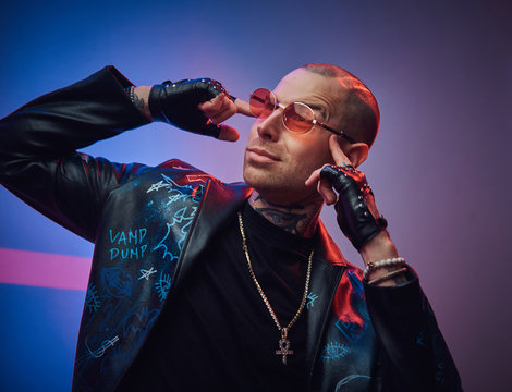 Creative, daring, tattooed, bald male rocker model posing in a studio for the photoshoot wearing fashionable designer black leather jacket, black shirt, red glasses, and jewelery, touching gead with