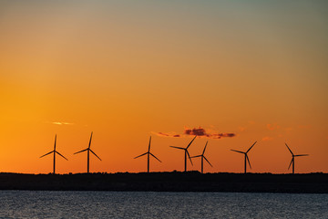 Evening sunlight on coast, pink and golden clouds and wind turbine. Sky reflection on water.  Wind generator for electricity, alternative energy source. Windmill for electric power production.
