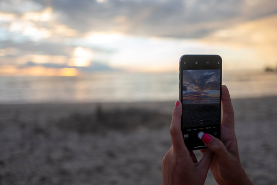 two hands using a cell phone to take a picture of the sunset at the beach