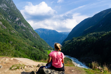 A woman and her child sit on a stone looking at a landscape with mountains and a mountain river on a sunny day with cloudy sky in Norway. The woman has blonde hair, and is dressed in a pink t-short.