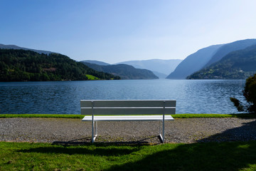 Naklejka na ściany i meble An empty white bench on shore of a Fjord in Norway with mountains in the background on a sunny day with bright sky.