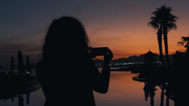 Woman taking photos of the sunset at luxury tropical resort. Girl at dusk takes pictures of beautiful summer sunset 4k