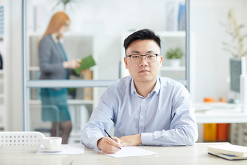 Fototapeta na wymiar Portrait of young Asian businessman wearing glasses and looking at camera while posing at workplace in office cubicle, copy space