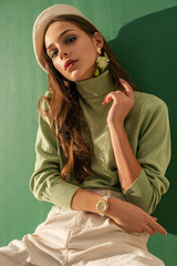 Young beautiful fashionable woman wearing green turtleneck, white beret, wrist watch, posing on mint color background. Spring fashion concept - obrazy, fototapety, plakaty