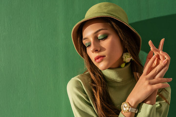 Close up portrait of young beautiful fashionable woman with green eyeshadow makeup, wearing turtleneck, trendy bucket hat, wrist watch, posing on mint color background. Copy, empty space for text - obrazy, fototapety, plakaty