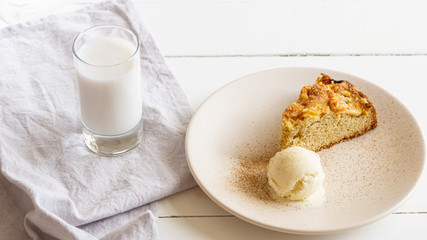 Apple pie slice with one bowl of vanilla ice cream on a round plate with some cinnamon in it. Near on a grey cotton napkin there is glass of milk. White wooden table. 