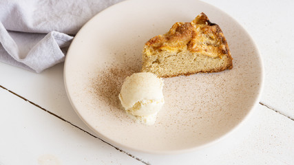 Apple pie slice with one bowl of vanilla ice cream on a round plate with some cinnamon in it. Near there is grey cotton napkin. White wooden table. 