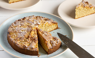 Sliced apple, cinnamon and almond pie on a blue round plate on a white wooden table. There is a knife near it and two plates with cake slices. 