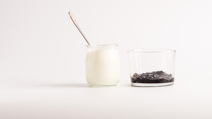 White natural yogurt in a glass jar with a spoon in it. Near there is a glass with some blueberry jam. White background. 