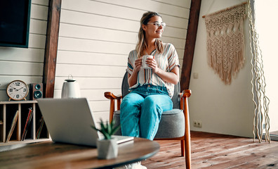 Enjoying time at home. Beautiful young smiling woman working on laptop and drinking coffee while sitting in a big comfortable chair at home.