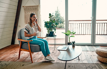 An attractive young woman in glasses is drinking coffee and talking on the phone while sitting in a comfortable chair at home.