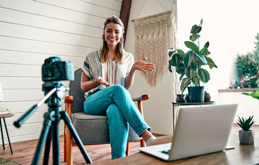 Attractive young woman blogger dressed in jeans and a blouse is recording on the camera while sitting on a comfortable chair at home. - Powered by Adobe