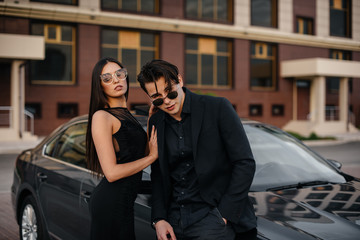 A young stylish couple in black stands near the car at sunset. Fashion and style