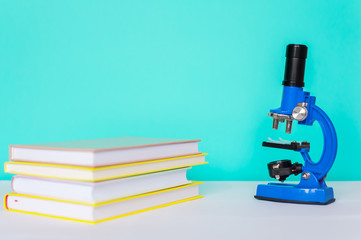 Microscope in blue on a green background with books on a white table. The concept of schooling, research and discovery.
