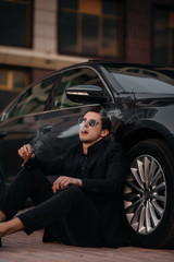 Fototapeta na wymiar A stylish businessman smokes cigars near a luxury car. Fashion and business