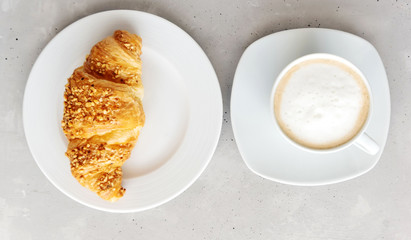 Classic French fresh croissant, sprinkled with nuts, on a white porcelain plate and a white mug of cappuccino, coffee with foam next to it. Gray background. View from above. Fresh breakfast.