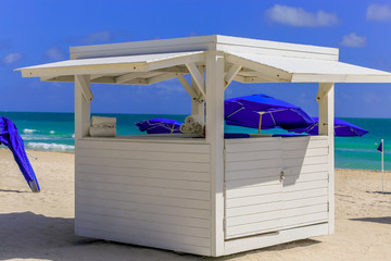 White pop up shed on beach supplying towels to guests at the beach with bright blue umbrellas, birght sunlight and small ocean waves.