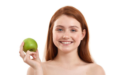 Only healthy food. Portrait of young and happy red-haired girl holding a fresh green apple and smiling while standing against white background