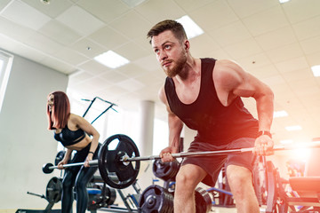 Muscular man and a young woman lifting heavy weights at gym. Fitness couple doing crossfit workout happily on the light modern background.