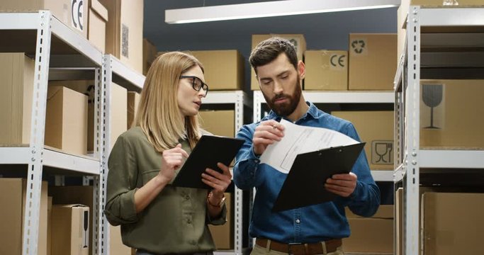Caucasian young couple of male and female postal workers standing in mail store with carton boxes, talking and registering parcels. Woman with tablet device tapping on and man filling in invoice.