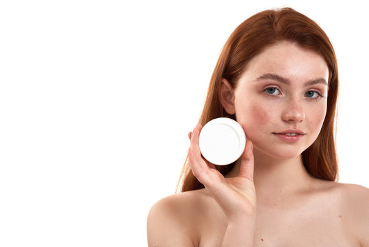 Perfect face cream for her skin type. Young attractive red-haired girl with freckles demonstrating cosmetic product and looking at camera while standing in studio against white background