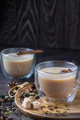 Traditional masala chai in glass cup with ingredients (cinnamon, anise, cardamom, nutmeg, pepper and brown sugar) on a dark stone background. 