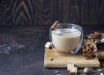 Traditional masala chai in glass cup with ingredients (cinnamon, anise, cardamom, nutmeg, pepper and brown sugar) on a dark stone background. 