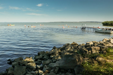 Easy afternoon on the shore of lake Balaton in Hungary. Rock on the bank in the foreground with swimmers and water activities in the background. A boat at the pier is seen .