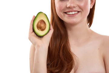Just healthy food. Cropped photo of young smiling red-haired girl holding a piece of avocado while standing against white background. Skin care