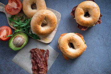 Bacon and egg bagel and salmon bagel on concrete background with avocado, tomato, rocket and bacon. Top view.