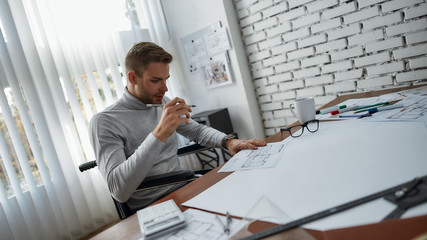 Inspiration. Young and handsome male engineer or architect in a wheelchair working with blueprint while sitting at his working place in the modern office