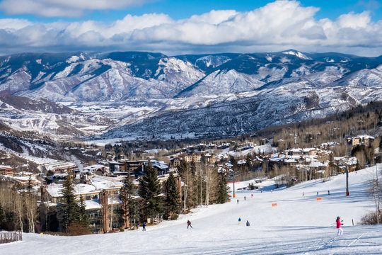 Panoramic view of Snowmass Village, with skiers skiing at the Aspen Snowmass ski resort in the foreground and the Rocky Mountains of Colorado in the background, on a partly cloudy winter day.  
