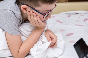 A preteen boy in glasses lying on a bed and watching video on smartphone