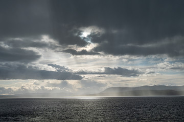 sunlight piercing through heavy cloud over the horizon illuminating the water surface on the ocean with mist of water surrounding the nearby island