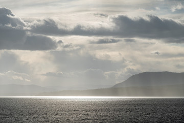 light piercing through heavy cloud over the horizon illuminating the water surface on the ocean with mist of water surrounding the nearby island